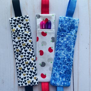 May include: Three fabric pen holders with different patterns. One has black and gold hearts on white, another has red apples and gray apples on white, and the third has a blue floral pattern. Each has a matching elastic loop.
