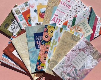 May include: A collection of rectangular paper booklets arranged in a fan shape, featuring diverse patterns including floral prints, cacti, postage stamps, hearts, and inspirational text such as "Enjoy Simple," "Hello," and "Difficult roads lead to beautiful destinations."