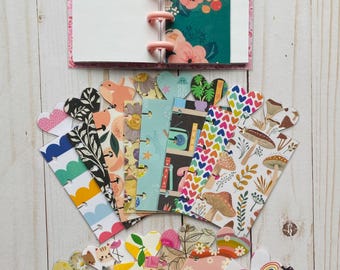 May include: A small disc-bound notebook with pink plastic discs and a floral page insert, accompanied by a collection of various patterned bookmarks featuring hearts, mushrooms, rainbows, cats, citrus fruits, and geometric designs.