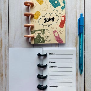 May include: A small notebook with a cream-colored cover featuring illustrations of telephones and the name "Riley" in a speech bubble. The notebook is open, revealing lined pages for contact information. A blue pen rests beside it.