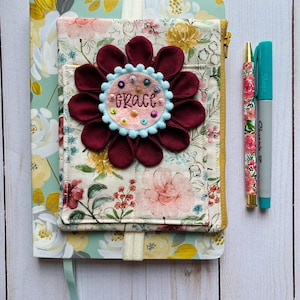 May include: A floral notebook with a matching fabric cover featuring a large maroon fabric flower with a light pink center embroidered with the name Grace and colorful beads. A floral patterned pen and a teal Sharpie marker are placed to the right.