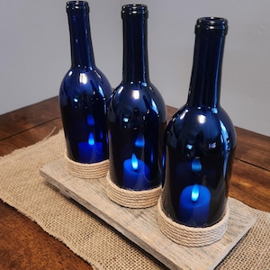 May include: Three dark blue glass bottles repurposed as candle holders. Each bottle has a rope accent around the base and holds a blue LED candle. The bottles are arranged on a wooden plank atop a burlap runner.