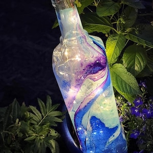 May include: A decorative glass bottle with a swirling blue, purple, and white design. The bottle is lit with small fairy lights and topped with a dark brown stopper. The bottle is set in a dark blue pot with greenery.