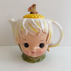 May include: A ceramic teapot shaped like a child's face with large black eyes and rosy cheeks. The lid features a yellow textured center resembling a flower with a small brown butterfly on top. The teapot has white and yellow petals as the rim.