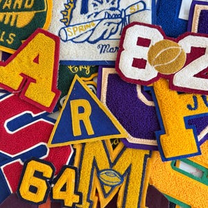 May include: A collection of assorted chenille varsity patches in various shapes and colors. Visible designs include a graduation cap, a basketball with the number 82, a triangle with the letter R, and a letter M featuring a football graphic.