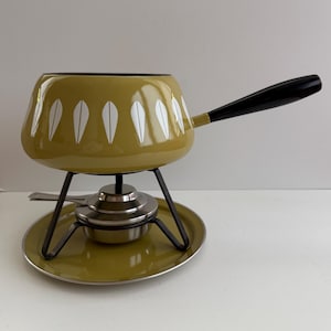 May include: An olive green fondue pot with a white leaf-like pattern, a long black handle, and a black metal stand. The set includes a stainless steel burner and a matching olive green circular base plate, creating a complete tabletop cooking unit.