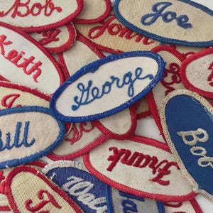 May include: A collection of vintage name patches in various colors. The patches are oval-shaped with embroidered names in red and blue thread. Some patches have a red border, while others have a blue border. The names include "Bob", "Joe", "Keith", and "George".