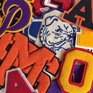 May include: A collection of colorful, textured varsity letter patches. The patches feature various letters, numbers, and school mascots in shades of orange, yellow, purple, red, and blue. The patches are made of felt and have a stitched border.