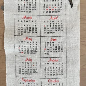 May include: A vintage calendar printed on a cream-colored fabric. The calendar displays the months from January to October, with each month's name in a decorative script. The days of the week and dates are printed in black.