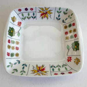 May include: A square white ceramic bowl featuring a decorative border of colorful floral illustrations. The pattern includes small red, yellow, and green flowers, leafy stems, and larger stylized yellow and red star-shaped blossoms.