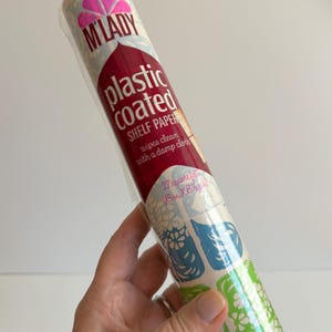 May include: A roll of M'Lady plastic coated shelf paper featuring a floral pattern in blue, green, and white. The packaging includes the text M'Lady, plastic coated, shelf paper, wipes clean with a damp cloth, and 27 square feet (18 in. X 18 yds.).