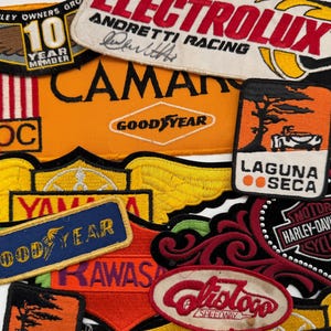 May include: A collection of embroidered patches featuring various logos and text. Notable patches include "ELECTROLUX ANDRETTI RACING", "CAMAN GOODYEAR", "LAGUNA SECA", and "HARLEY-DAVIDSON MOTOR CYCLES". The patches are in various colors, including orange, red, and black.