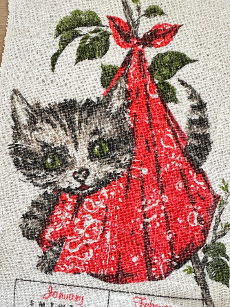 May include: Vintage fabric featuring a tabby kitten with green eyes, nestled in a red bandana tied to a branch with green leaves. The fabric has a calendar with the month of January visible.