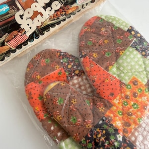 May include: Deluxe Cook & Bake MITTS oven mitts in a clear plastic package. The mitts have a patchwork design with brown, orange, green, and black floral and checkered patterns. The packaging reads "Palms coated with Teflon."