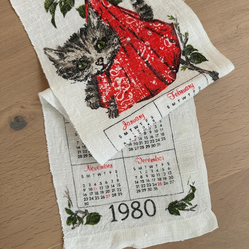 May include: A vintage linen calendar towel featuring a raccoon in a red outfit. The towel includes the months of January, February, November, and December, along with the year 1980. The design is printed in black, red, and green on a white background.