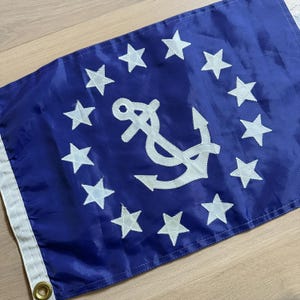 May include: A blue flag featuring a white anchor in the center, surrounded by a circle of white stars. The flag has a white border and a brass grommet. Nautical themed flag.