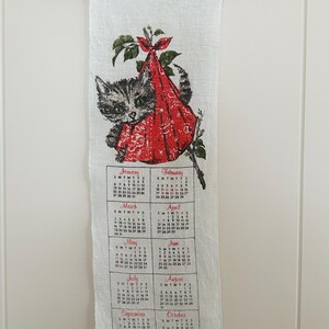 May include: A vintage 1980 calendar towel featuring a gray tabby cat wearing a red bonnet. The calendar displays months from January to December. The towel is hanging from a red and green crochet hanger.