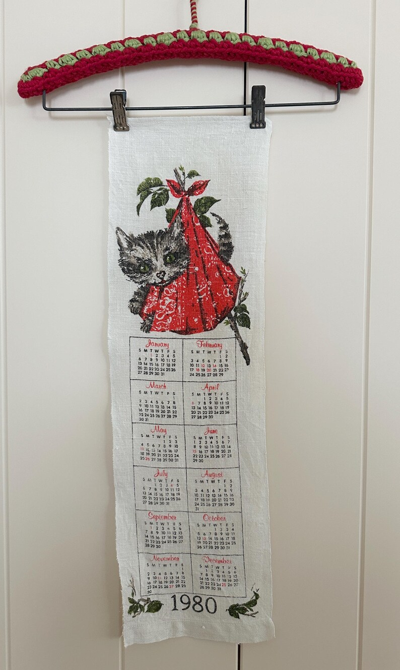 May include: A vintage 1980 calendar towel featuring a gray tabby cat wearing a red bonnet. The calendar displays months from January to December. The towel is hanging from a red and green crochet hanger.