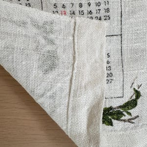 May include: Close-up of a linen calendar for October. The calendar is printed in black and red ink, with the month name at the top. Green botanical illustrations are visible. The fabric is folded, revealing the texture.