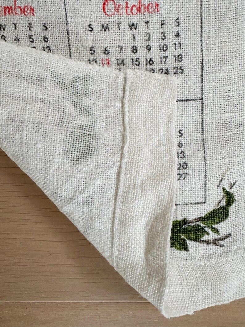 May include: Close-up of a linen calendar for October. The calendar is printed in black and red ink, with the month name at the top. Green botanical illustrations are visible. The fabric is folded, revealing the texture.
