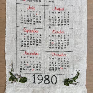 May include: A vintage linen calendar towel featuring the year 1980. The calendar displays months from July to December, with each month's days and dates printed in black and red. Green floral accents are at the bottom.