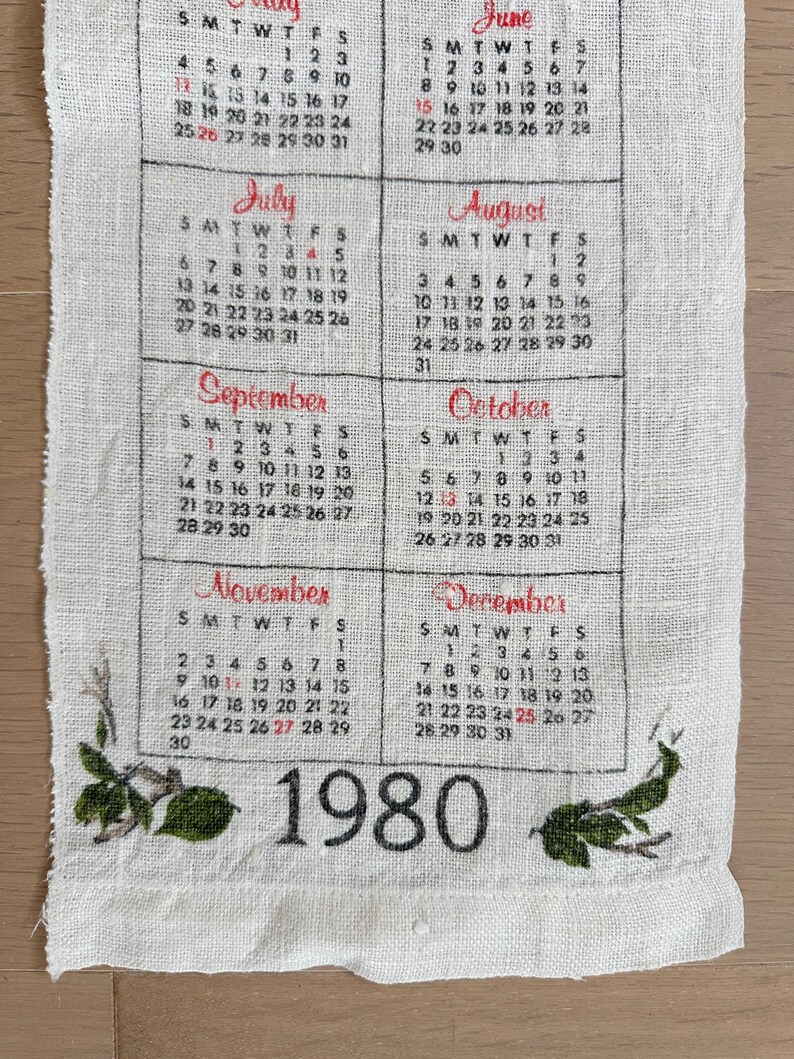 May include: A vintage linen calendar towel featuring the year 1980. The calendar displays months from July to December, with each month's days and dates printed in black and red. Green floral accents are at the bottom.
