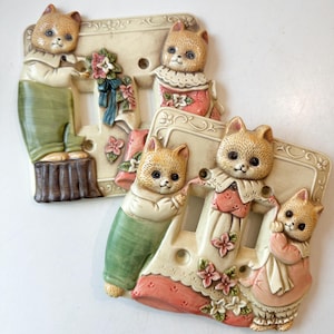 May include: Two decorative light switch covers featuring a whimsical design. Each cover has a rectangular shape with a cream-colored background and a detailed relief of a cat dressed in vintage clothing. The cats are holding flowers and are surrounded by floral accents.