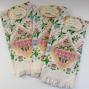 May include: Three off-white cotton kitchen towels with fringed edges, featuring a diamond grid pattern, floral motifs in pink, green, and blue, and a central pink diamond containing the text COUNTRY HOME. Each towel has a Homespun Hearth tag.