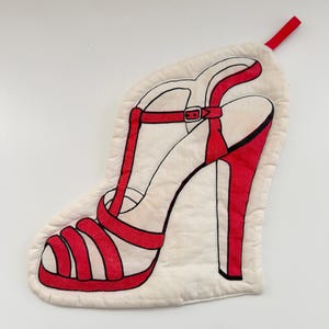 May include: A white oven mitt shaped like a high-heeled shoe. The shoe design features red straps, a heel, and a buckle. A red loop is attached at the top for hanging. The mitt is made of a quilted fabric.