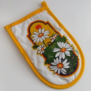 May include: White oven mitt with a yellow border and hanging loop. The mitt features a floral design with large white daisies, green leaves, and a yellow background with a red sun and a small house.