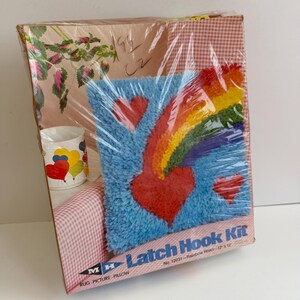May include: A square latch hook kit in a plastic-wrapped box featuring a blue background with a rainbow and red hearts. The box displays the text Latch Hook Kit, No. 12031-Rainbow Heart-12 x 12, and MH logo. A white mug with heart graphics is also visible.