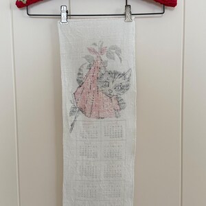 May include: A vintage white linen calendar towel featuring a cat and floral design, hanging from a red and green crocheted hanger. The calendar is printed in black ink, with the year 1980 visible at the bottom.