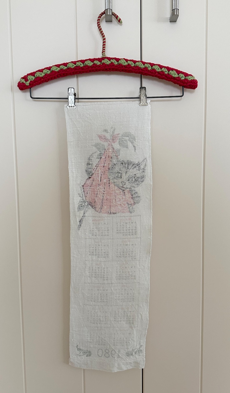 May include: A vintage white linen calendar towel featuring a cat and floral design, hanging from a red and green crocheted hanger. The calendar is printed in black ink, with the year 1980 visible at the bottom.