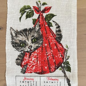 May include: A vintage linen calendar towel featuring a gray tabby kitten peeking out of a red patterned sack. The towel includes the months of January, February, March, and April with their respective calendar dates. Green leaves and branches frame the kitten.