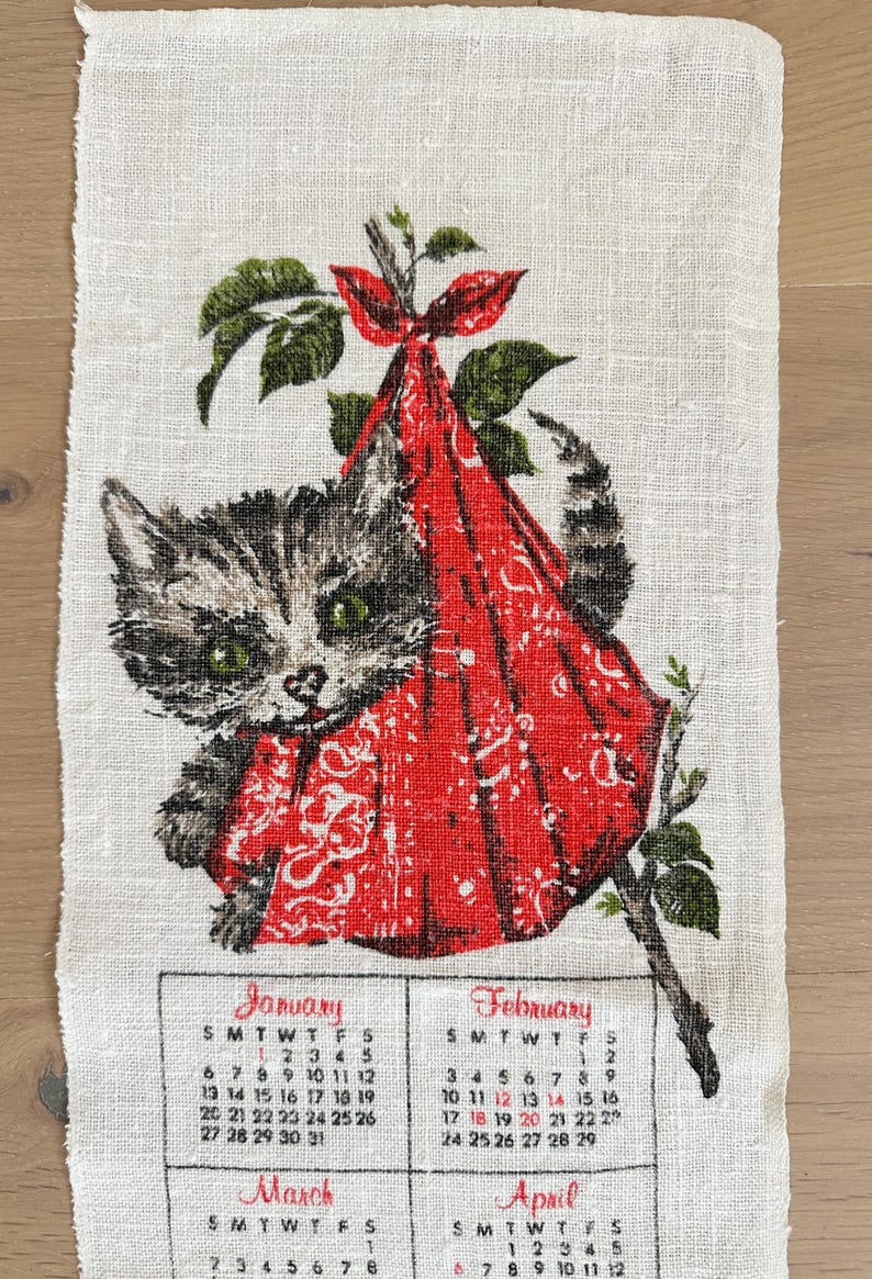 May include: A vintage linen calendar towel featuring a gray tabby kitten peeking out of a red patterned sack. The towel includes the months of January, February, March, and April with their respective calendar dates. Green leaves and branches frame the kitten.
