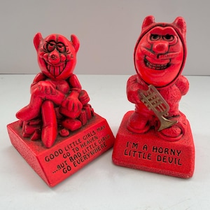May include: Two red devil figurines on rectangular bases. The left figure sits with legs crossed, featuring the text: GOOD LITTLE GIRLS MAY GO TO HEAVEN... BUT BAD LITTLE GIRLS GO EVERYWHERE. The right figure holds a trumpet, with the text: I'M A HORNY LITTLE DEVIL.