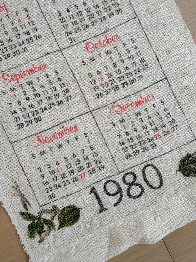 May include: Vintage 1980 calendar printed on a linen-like fabric. The calendar features months like September, October, November, and December, with dates in black and red. Floral accents are visible at the edges.