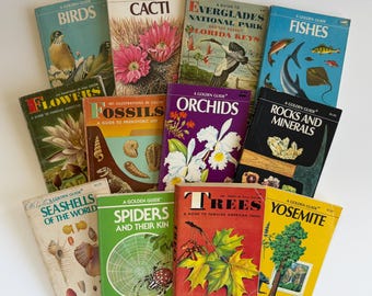 May include: A collection of vintage Golden Guide books, featuring titles like "Birds," "Cacti," "Fossils," and "Spiders and Their Kin." The books have colorful illustrations and are arranged on a white surface. The books are a mix of colors, including blue, green, yellow, and purple.