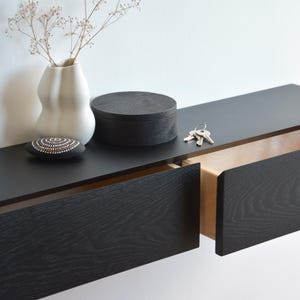 May include: A black floating shelf with two drawers. The top drawer is open and the bottom drawer is closed. The shelf has a natural wood interior. The shelf is mounted on a white wall.