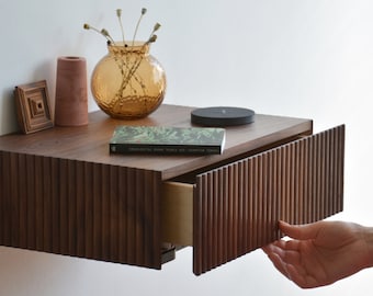 Puede incluir: Una mesita de noche flotante de madera marrón oscuro con un cajón abierto. El cajón tiene un diseño acanalado. La mesita de noche tiene un libro, una plataforma de carga inalámbrica y un jarrón con flores secas en la parte superior.