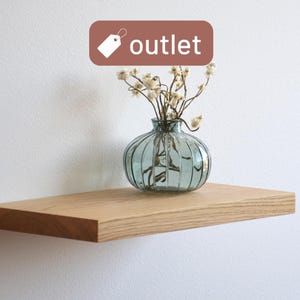 Op de afbeelding: Een licht houten zwevend plankje met een heldere, geribbelde glazen vaas met gedroogde bloemen. Het plankje is aan een witte muur bevestigd. Een bruin label met het woord "outlet" staat in de linkerbovenhoek.