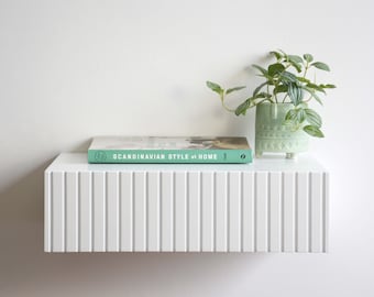 May include: A white floating shelf with a book titled "Scandinavian Style at Home" and a small green potted plant on top. The shelf has a grooved design.