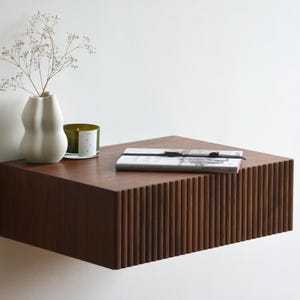 May include: A dark brown wooden floating shelf with a fluted front. The shelf is decorated with a white vase with dried flowers, a glass candle holder with a green candle, and a book with a black ribbon bookmark.