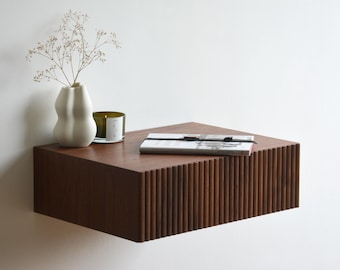 Puede incluir: Una estantería flotante de madera marrón oscuro con un frente acanalado. La estantería está decorada con un jarrón blanco con flores secas, un portavelas de vidrio con una vela verde y un libro con un marcapáginas de cinta negra.