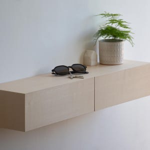 Puede incluir: Una estantería flotante de madera clara con dos cajones, unas gafas de sol y una pequeña planta en maceta en la parte superior.