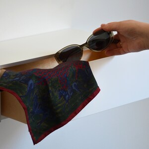 May include: A white shelf with a brown drawer that is partially open. A patterned fabric handkerchief is pulled out of the drawer. A pair of sunglasses is being placed on top of the shelf.