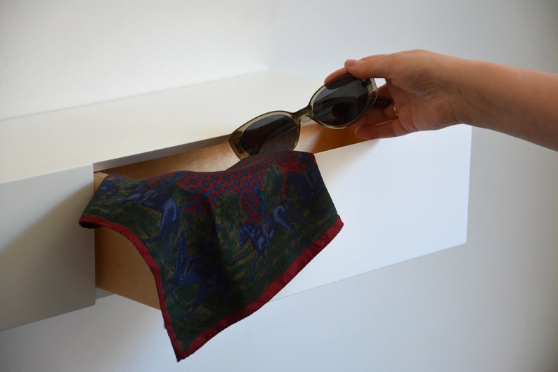 May include: A white shelf with a brown drawer that is partially open. A patterned fabric handkerchief is pulled out of the drawer. A pair of sunglasses is being placed on top of the shelf.