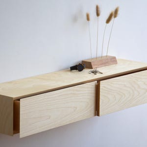 Könnte beinhalten: Helles, schwebendes Holzregal mit zwei Schubladen, an einer weißen Wand befestigt. Das Regal ist mit einem kleinen Holzblock mit getrockneten Pflanzen, einer schwarzen Uhr und einem Schlüsselbund dekoriert. Das Regal ist etwa 100 cm lang.