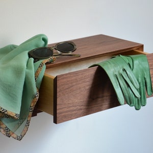 May include: A wall-mounted wooden drawer unit with a dark wood grain finish. A light green scarf with a plaid border and dark-rimmed sunglasses rest on top, while a pair of green leather gloves hangs over the edge of the open drawer.