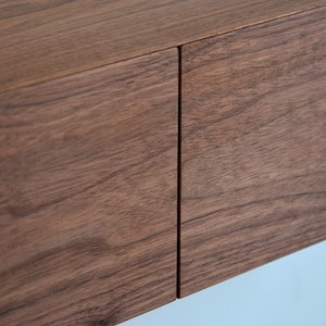 May include: A close-up view of two rectangular wooden drawer fronts featuring a rich, dark brown grain pattern. The panels are positioned side-by-side with a thin, vertical gap separating them, showcasing the natural texture and horizontal wood grain.